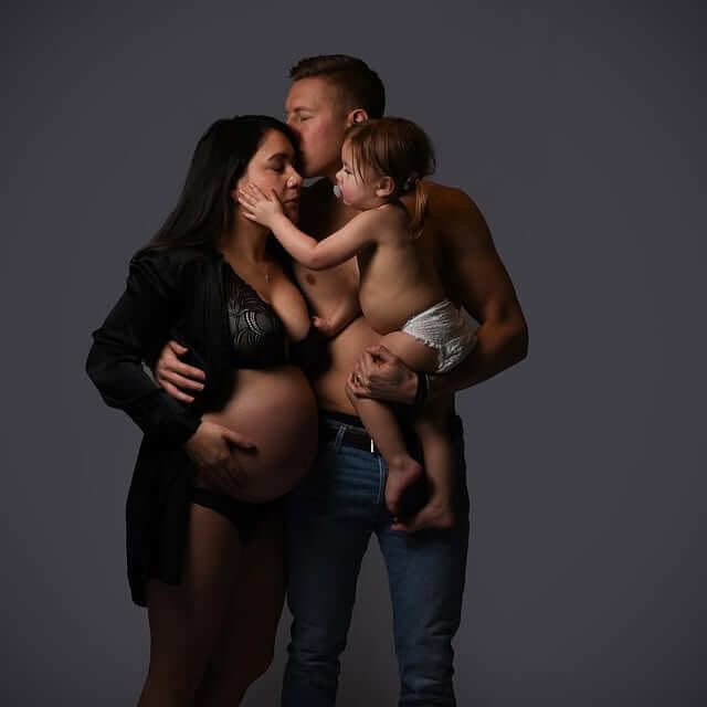 Familjeporträtt av ett par med ett litet barn, där barnet håller om mammans ansikte och pappan kysser hennes panna