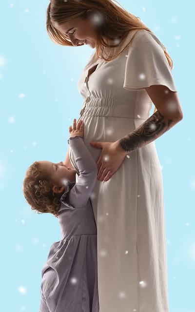Litet barn håller om sin gravida mamma i vit klänning, med vintereffekt i bakgrunden.