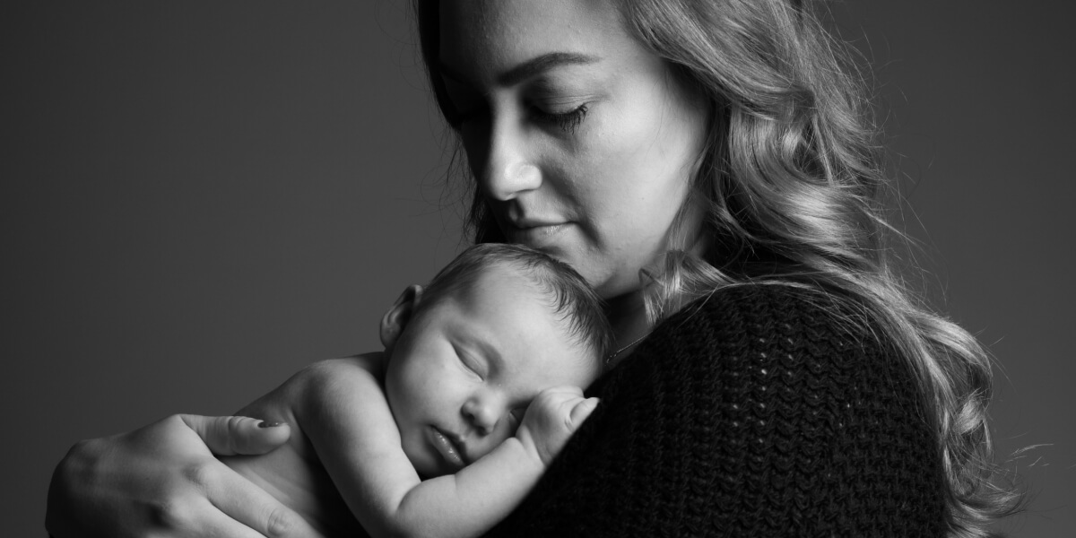 Svartvit bild på en mamma som håller sin nyfödda bebis tätt intill bröstet med ömhet i blicken